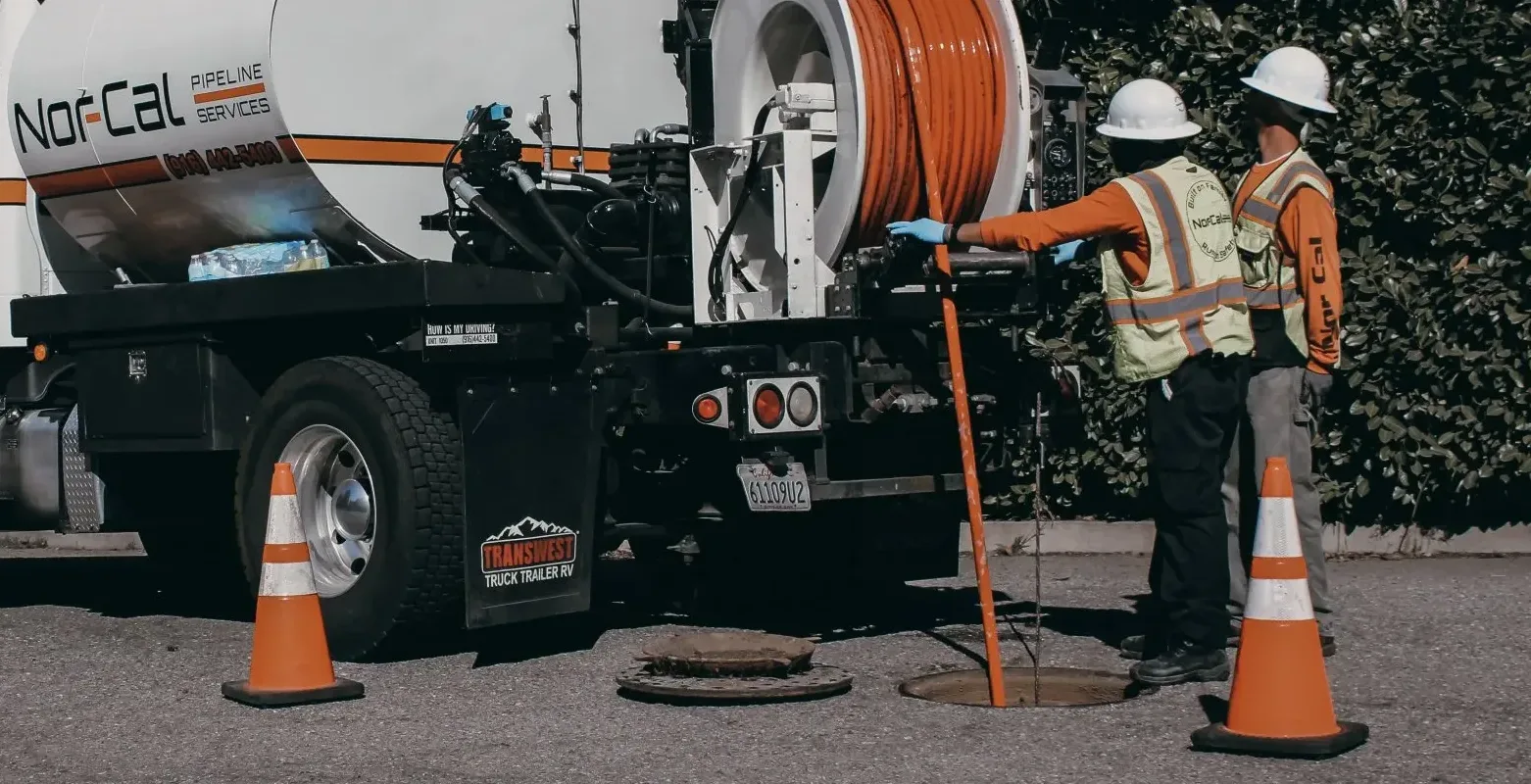Two workers in safety gear operating a Nor-Cal Pipeline Services truck with high-pressure water jetting equipment near a manhole, emphasizing pipe jetting services for clearing blockages and restoring flow.
