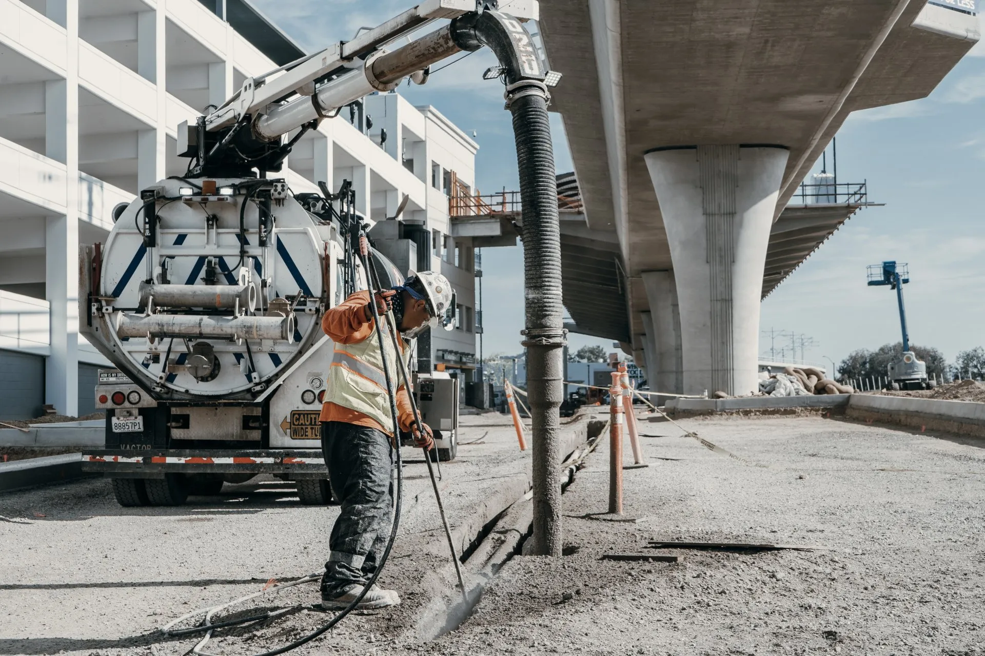 Hydro Excavation Worker operating hydro excavation equipment on construction site, with utility truck and elevated concrete structure in background, showcasing Nor-Cal Pipeline Services' underground utility services.
